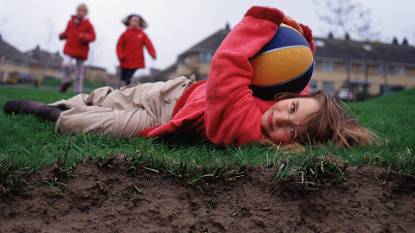 Meisje met bal ligt op gras waarbij aarde onder gras zichtbaar is, twee andere meisjes komen aanrennen