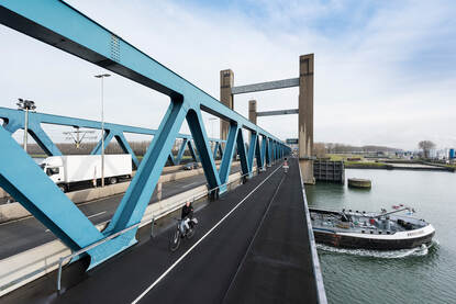 Foto van fietser en vrachtauto op brug waar boot onderdoor vaart